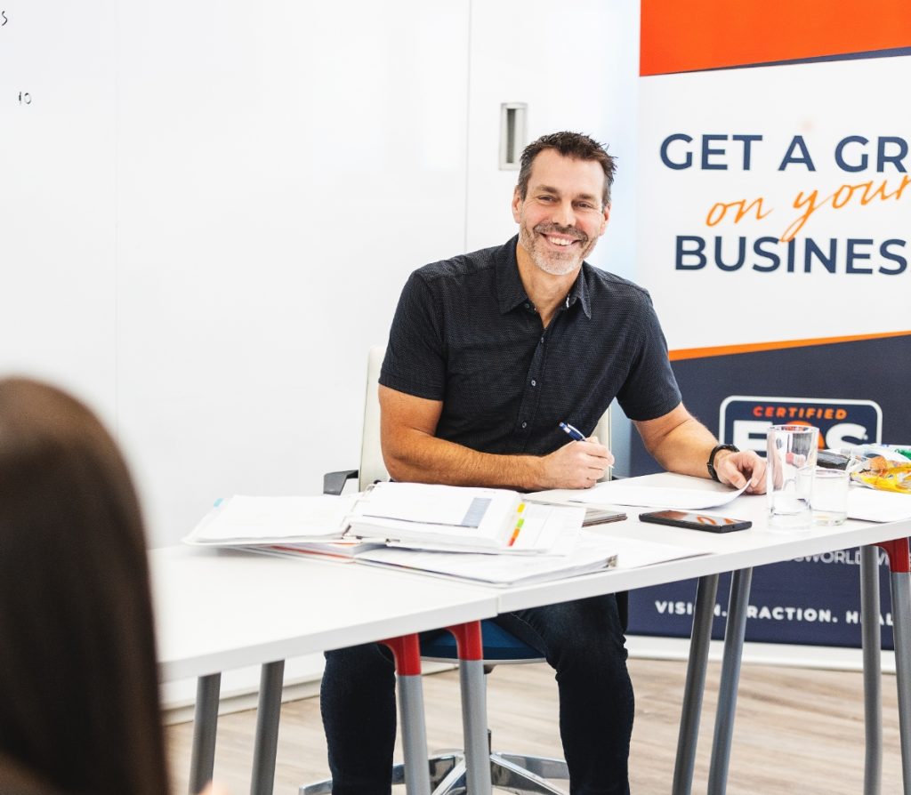 Jon Weening smiling while sitting at a table near EOS signage